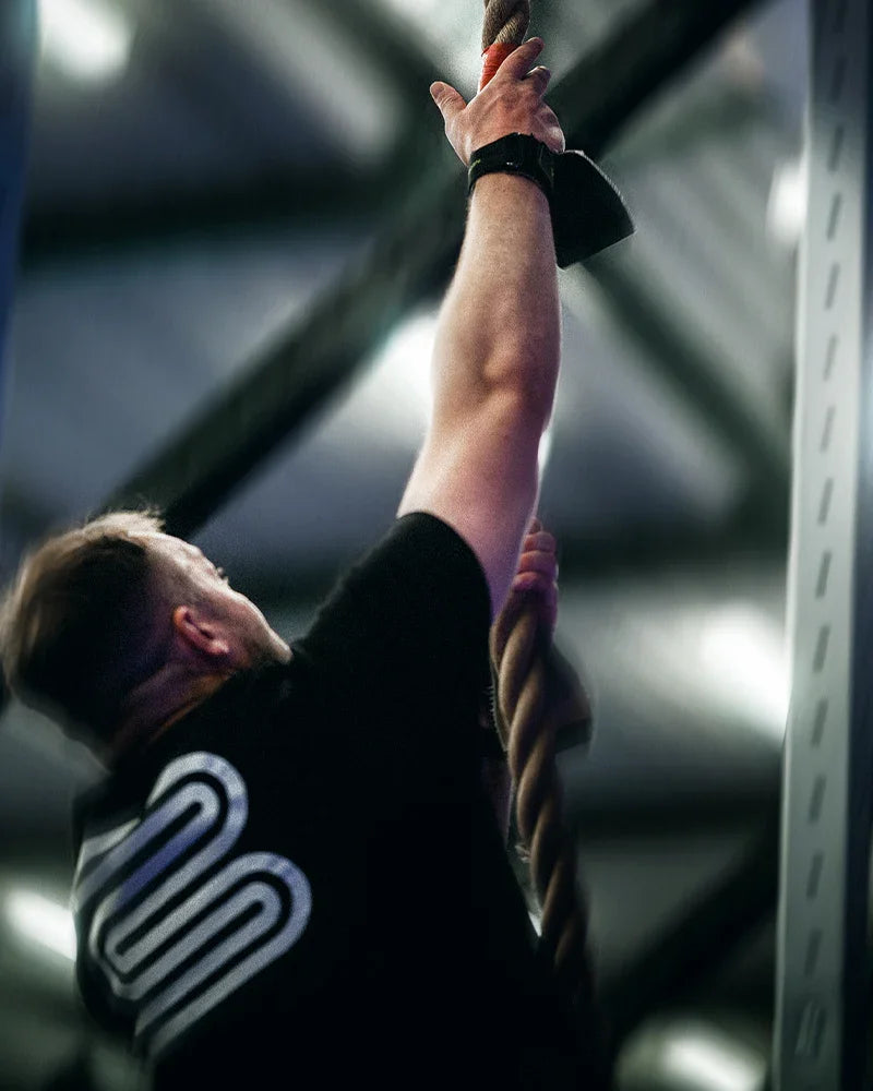 Person wearing black shirt with white logo climbing a rope in a gym setting